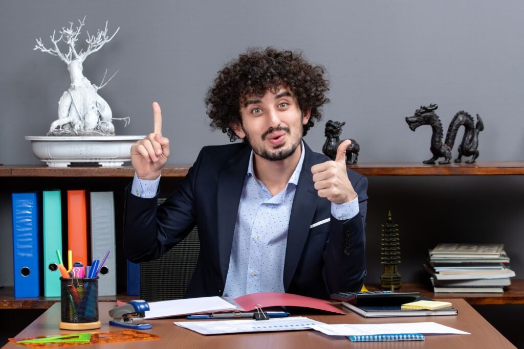 Cheerful professional sitting at office desk, pointing upward and giving thumbs up, surrounded by files, stationery, and decorative objects.
