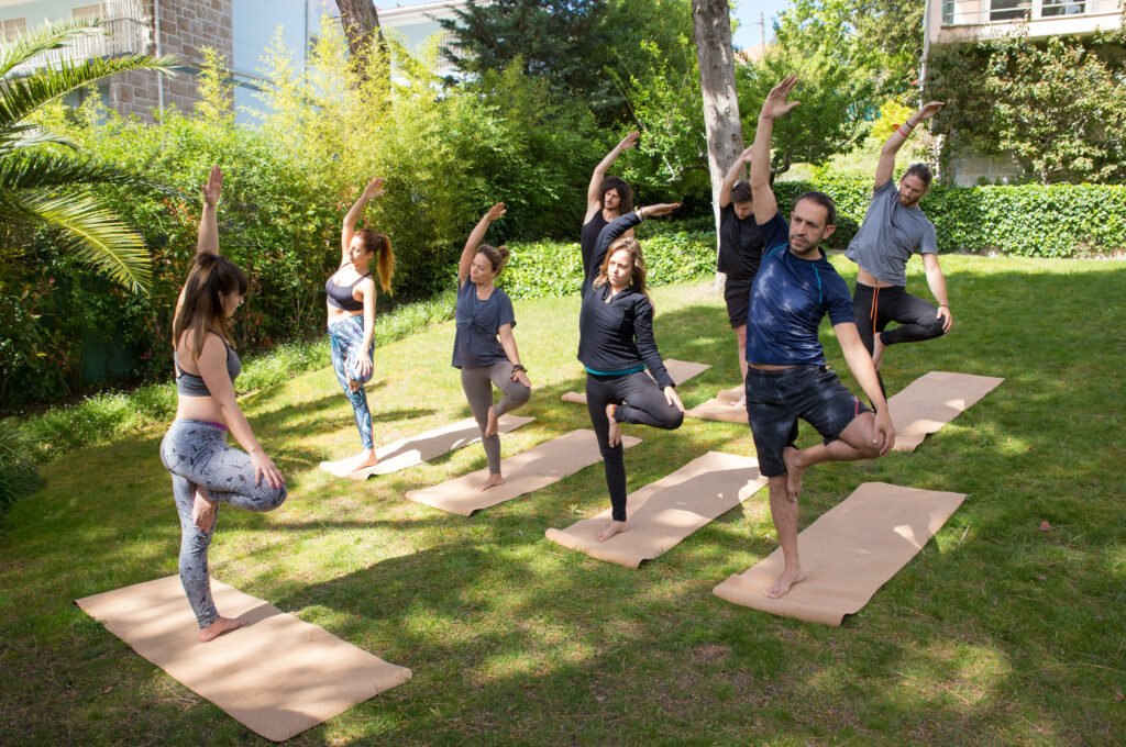 Group of people practicing yoga outdoors on mats, performing balance poses in a sunny, green garden setting.