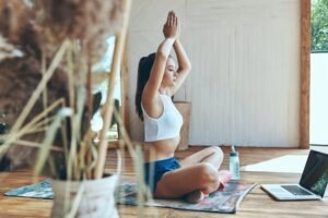 Woman practicing seated yoga with raised arms on a mat indoors, laptop nearby, focusing on mindfulness, balance, and breathing.