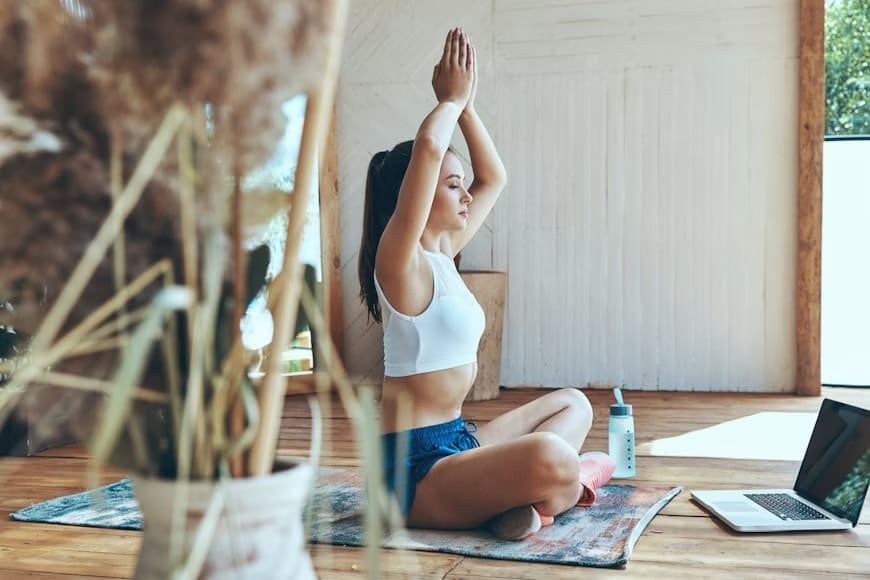 Woman practicing seated yoga with raised arms on a mat indoors, laptop nearby, focusing on mindfulness, balance, and breathing.