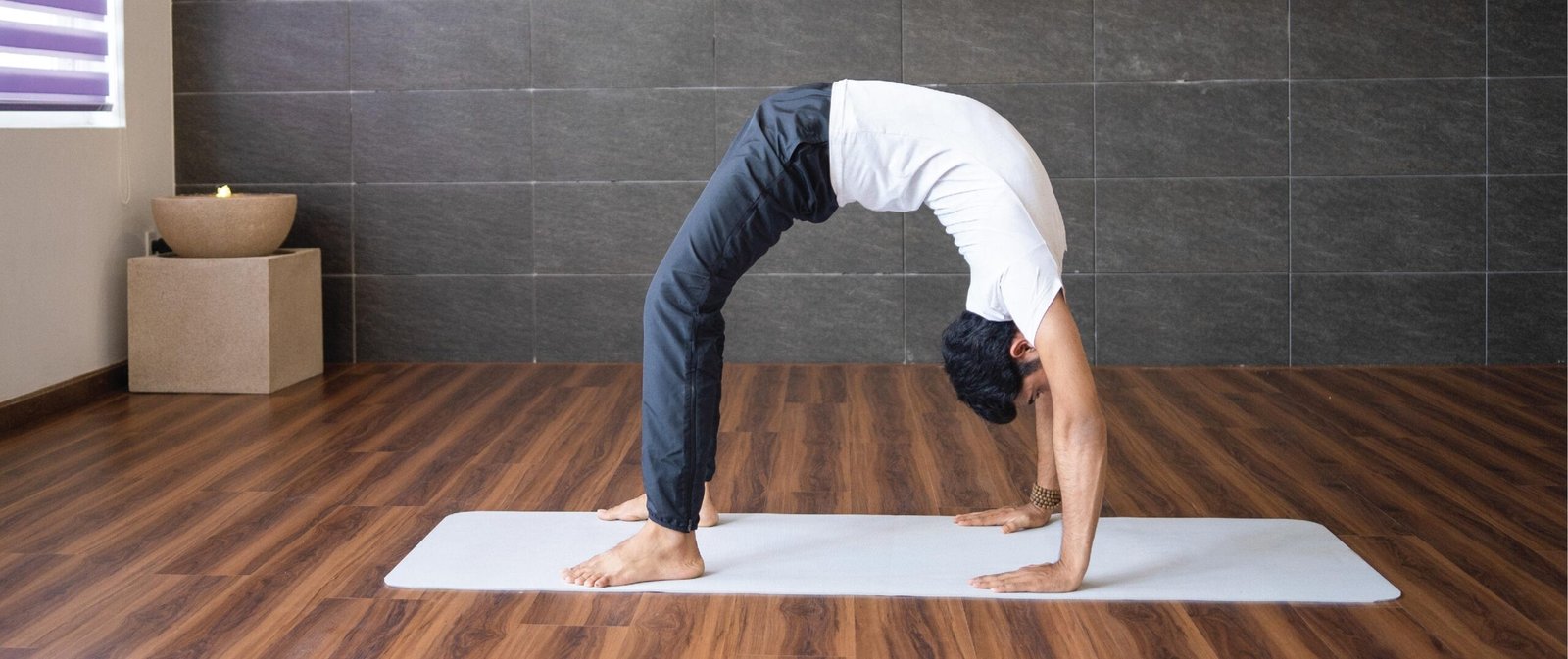 Person performs wheel pose yoga posture on a mat indoors, arching back with hands and feet grounded on wooden floor.