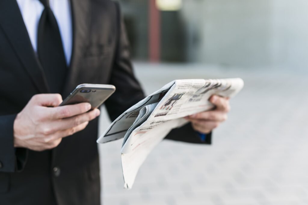 Business professional holding a smartphone and newspaper simultaneously, checking digital updates while reading printed news outside an office building.