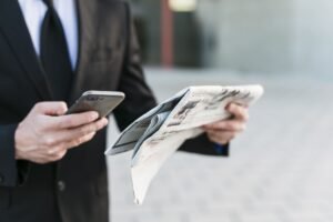 Business professional holding a smartphone and newspaper simultaneously, checking digital updates while reading printed news outside an office building.