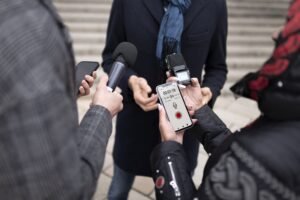 Reporters holding microphones and smartphones interview a spokesperson outdoors, recording audio and notes during a media interaction on public steps.