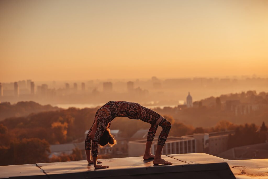 Person performing advanced yoga backbend on rooftop at sunrise, overlooking misty city skyline, balanced posture emphasizing strength, flexibility, and focus.