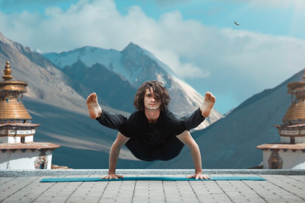 Person performing advanced arm balance yoga pose outdoors, mountains and temple structures behind, demonstrating strength, control, focus, and mindful practice.